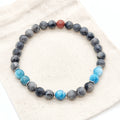 A beaded stretch bracelet featuring dark gray and black labradorite beads, bright blue apatite beads, and a single red jasper accent bead, displayed on a beige textured fabric background.