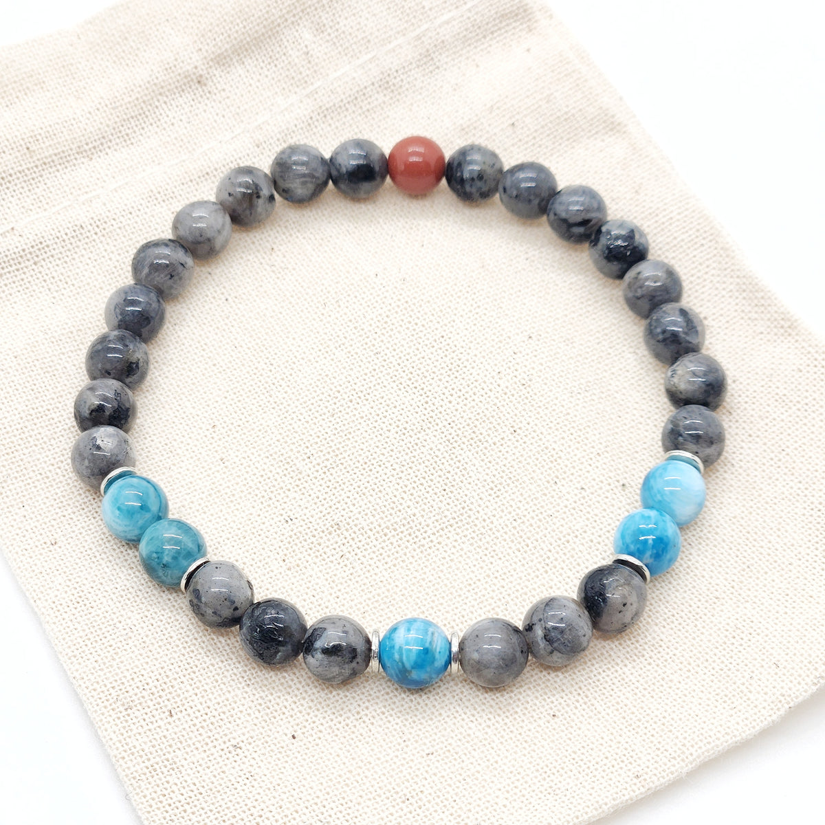 A beaded stretch bracelet featuring dark gray and black labradorite beads, bright blue apatite beads, and a single red jasper accent bead, displayed on a beige textured fabric background.