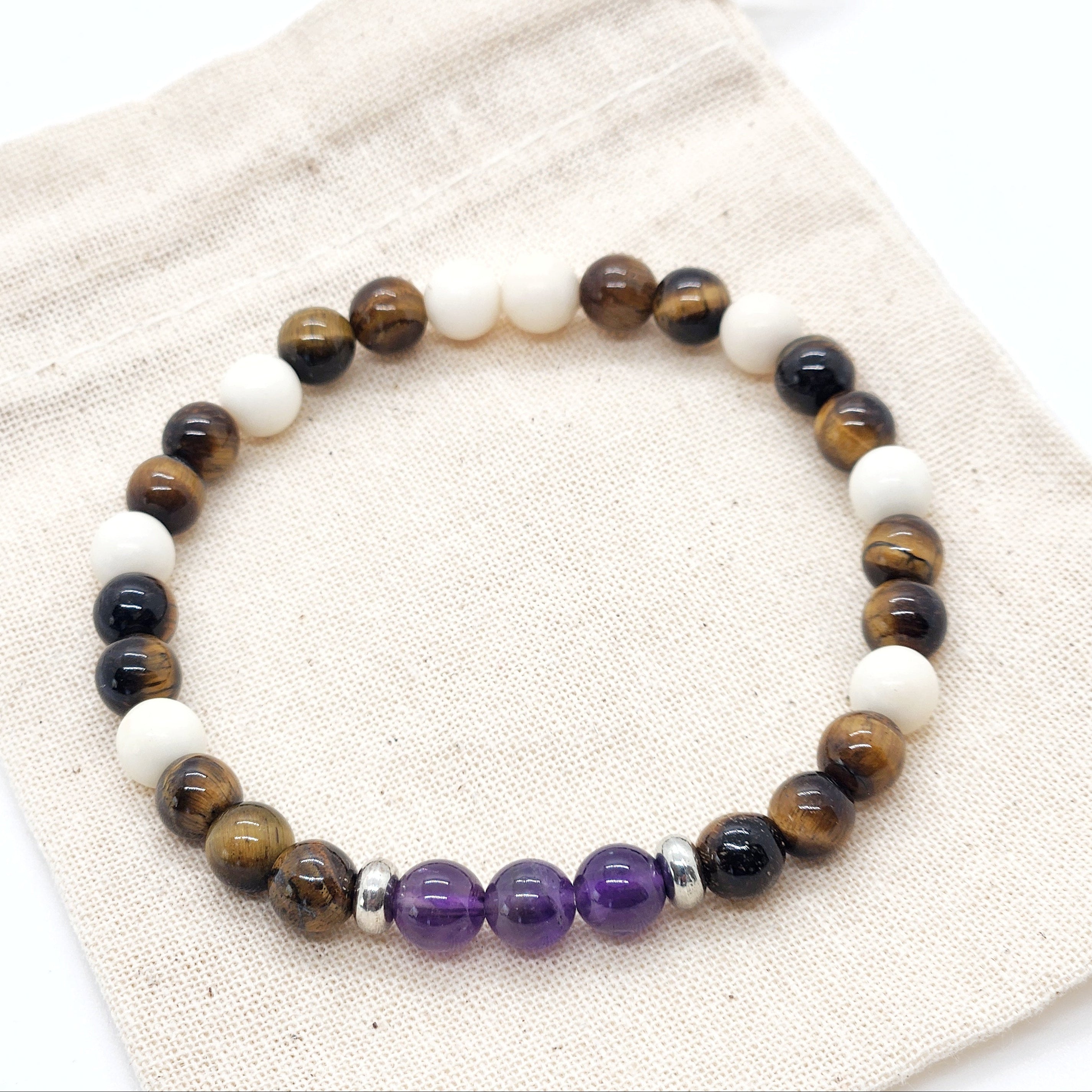 A beaded stretch bracelet made of brown tiger's eye, cream jasper beads, and purple amethyst beads, displayed on a beige canvas drawstring bag.