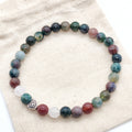 A beaded stretch bracelet made of round multicolored gemstone beads, featuring a small silver Celtic knot heart charm, displayed on a textured beige fabric background.