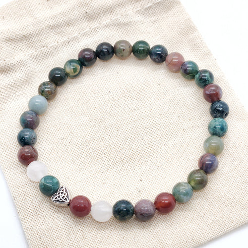 A beaded stretch bracelet made of round multicolored gemstone beads, featuring a small silver Celtic knot heart charm, displayed on a textured beige fabric background.
